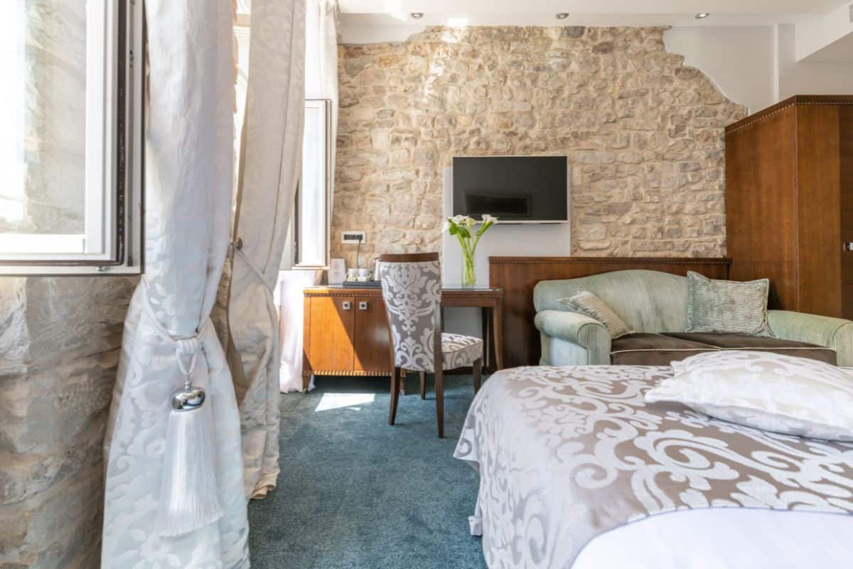 A room at Heritage Hotel Antique Split, one of the top boutique hotels in Croatia, featuring a king bed with damask bedding, exposed stone wall, teal carpet, and traditional dark wood furniture.