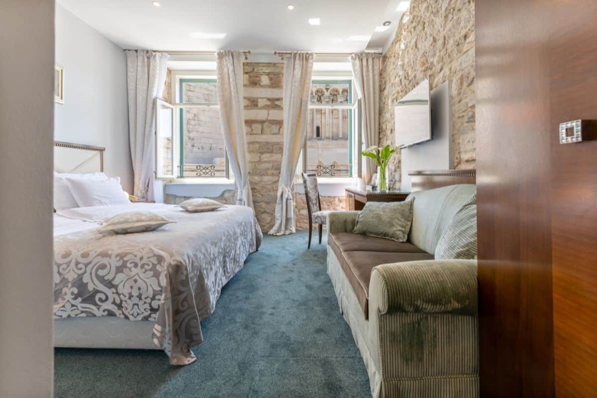 A bright room at Heritage Hotel Antique Split featuring a king bed, green sofa, exposed stone wall, and open windows with views of a historic building in Split's old town.