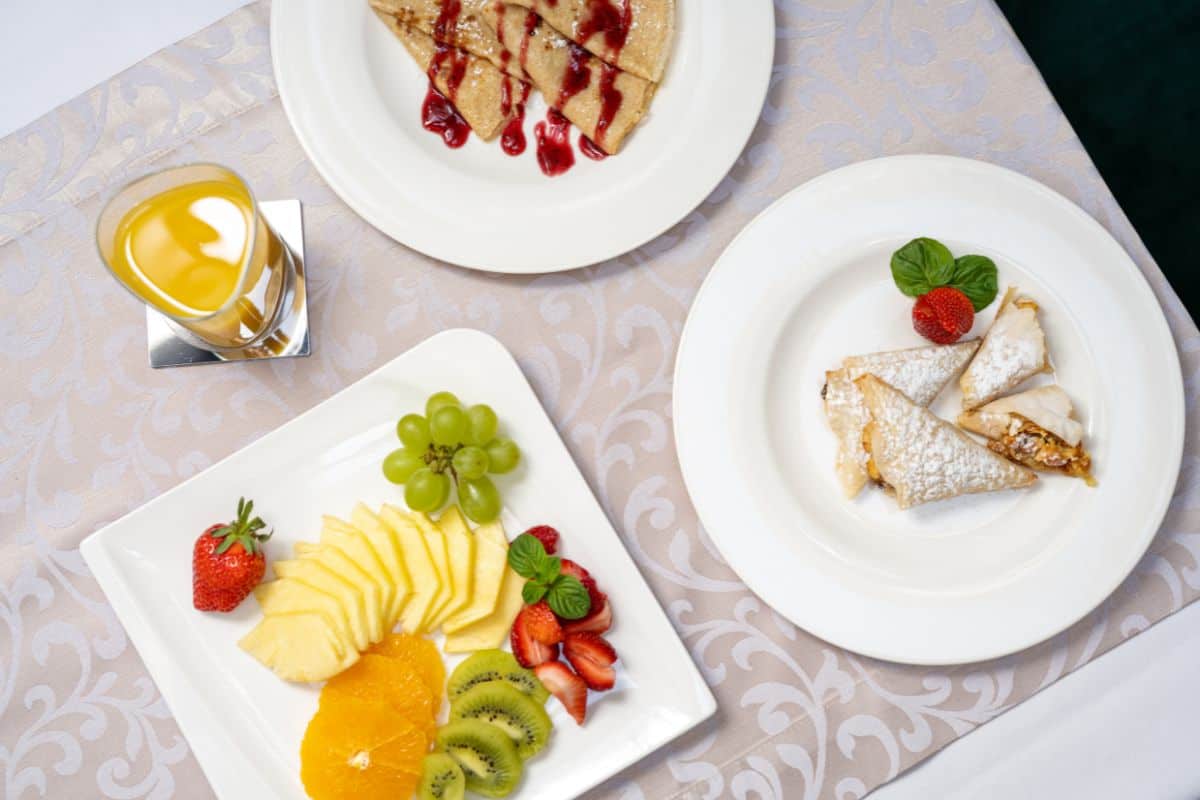 A breakfast spread at Heritage Hotel Antique Split with crepes drizzled in berry sauce, a fresh fruit platter of pineapple, strawberries, kiwi and grapes, and a glass of orange juice.