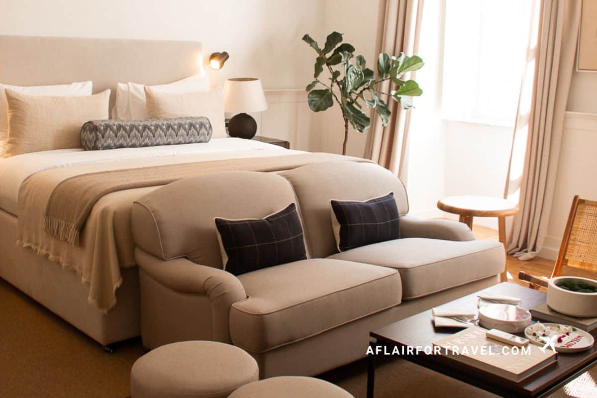 A serene and modern room at Boutique Hotel Stari Grad Dubrovnik, featuring a king bed with neutral linen bedding, a cream sofa with navy cushions, fiddle leaf fig plant, and warm natural light.