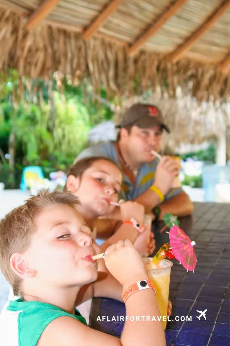 A boy, a girl, and their dad sip tropical drinks under a palm-thatched pool bar
