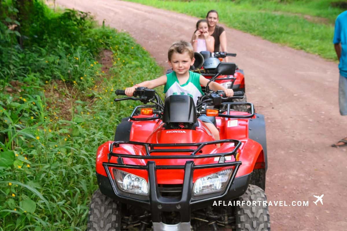 A boy sitting on an ATV along a dirt trail, with his mom and sister close behind