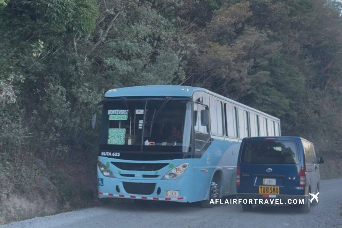 A blue public bus on Route 625 navigating a narrow, tree-lined mountain road alongside a tourism van which is what to expect in Monteverde, Costa Rica when driving there.