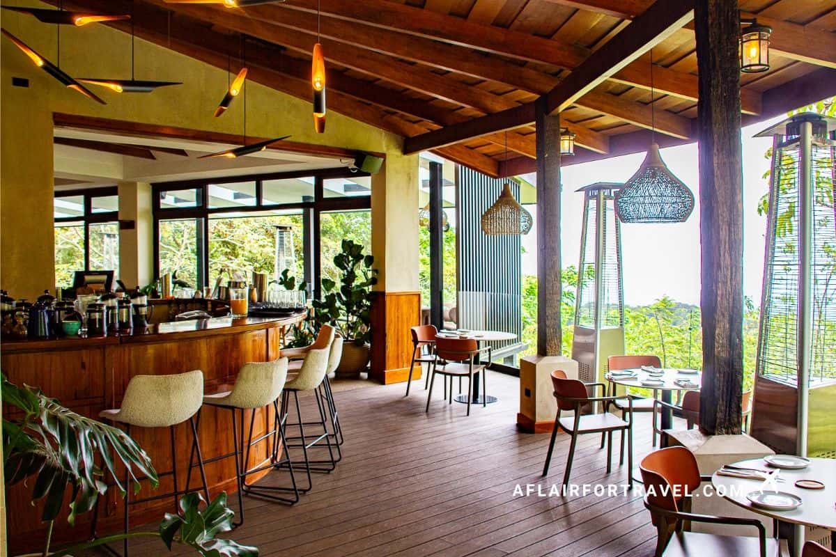 An open-air hotel restaurant with a wooden bar, wicker pendant lights, and floor-to-ceiling views of lush Costa Rican forest