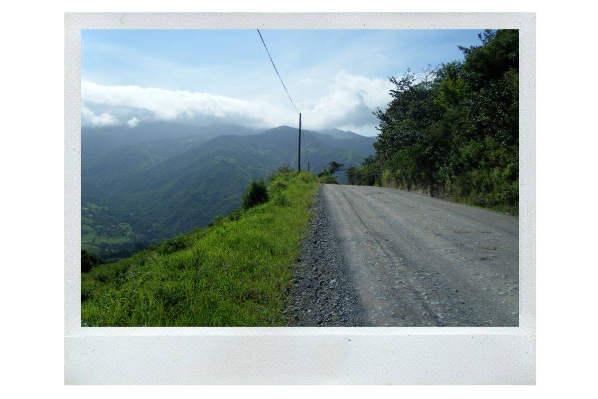 A narrow gravel road winding along a steep hillside with sweeping views of green mountains and low clouds in Costa Rica