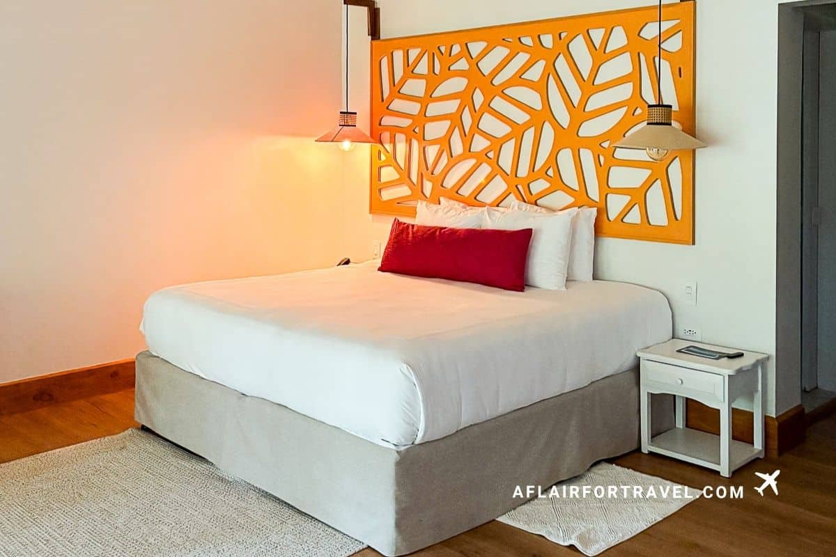 A bright hotel room with a white king bed, red accent pillow, and an orange leaf-pattern carved wooden headboard