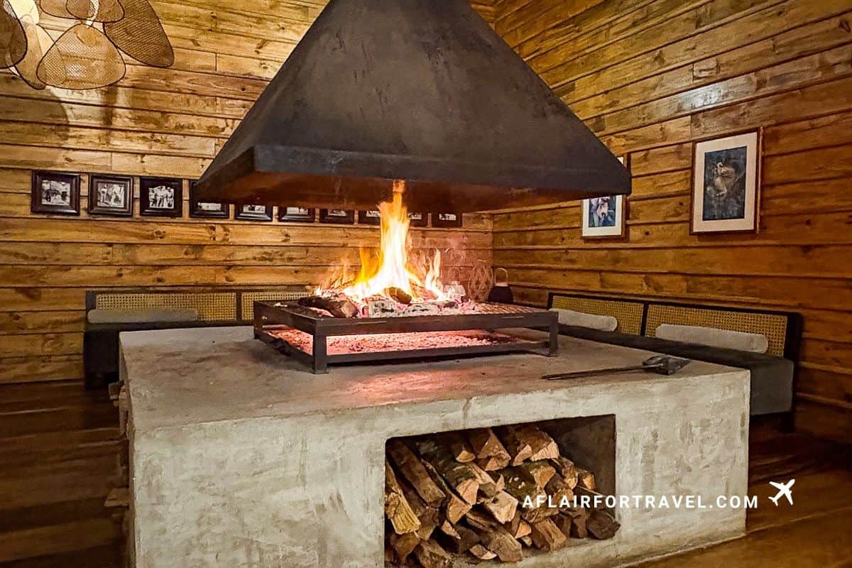 A rustic wood-paneled lodge dining room with a large central open fire pit surrounded by bench seating