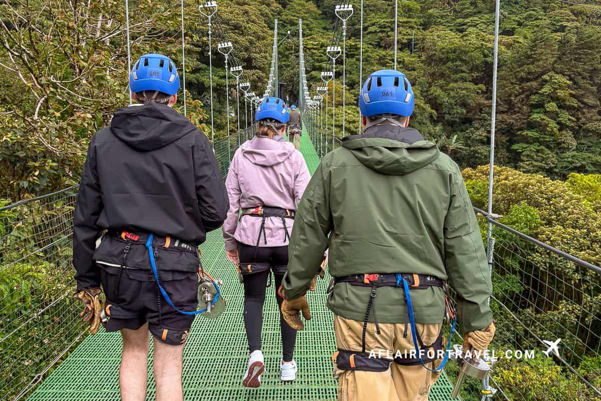 Three visitors in helmets and safety harnesses walking across a green suspended canopy bridge above the cloud forest which is what to expect in Monteverde, Costa Rica.