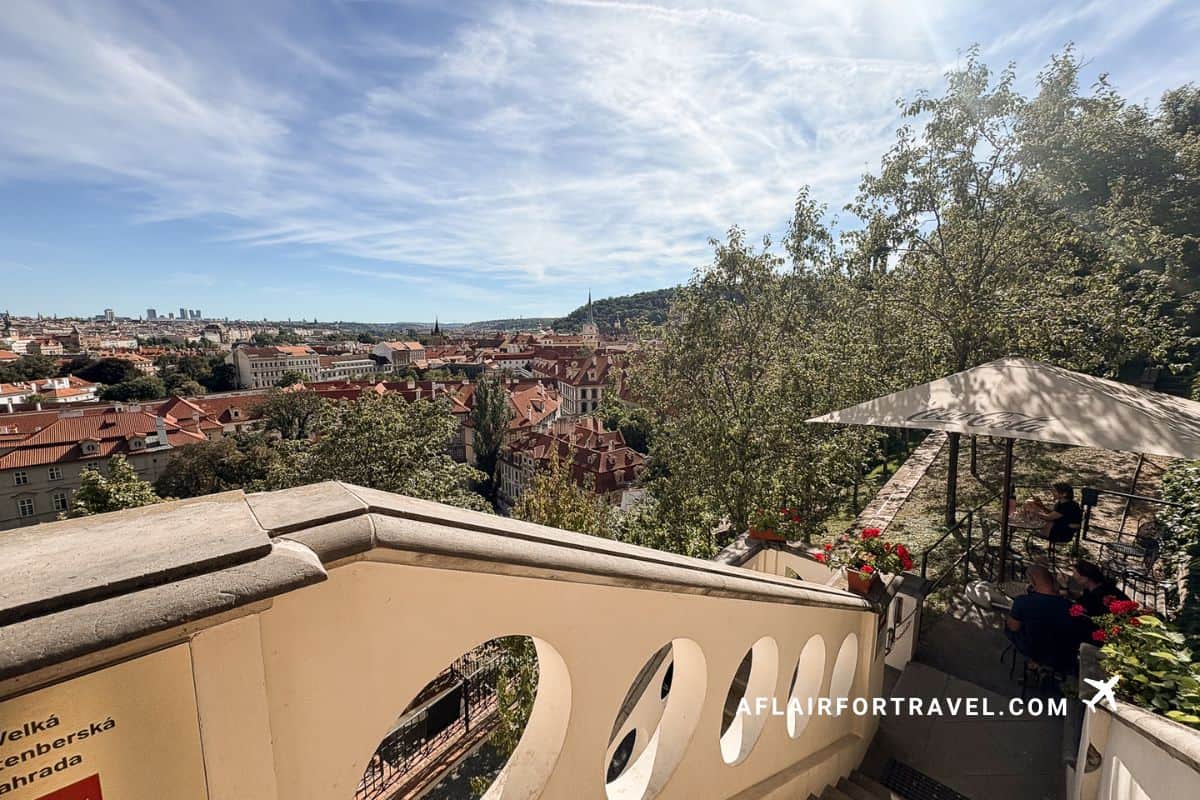 View over Prague's red rooftops from the terrace of Vrtbovská Garden, with castle hill and a shaded café in the background
