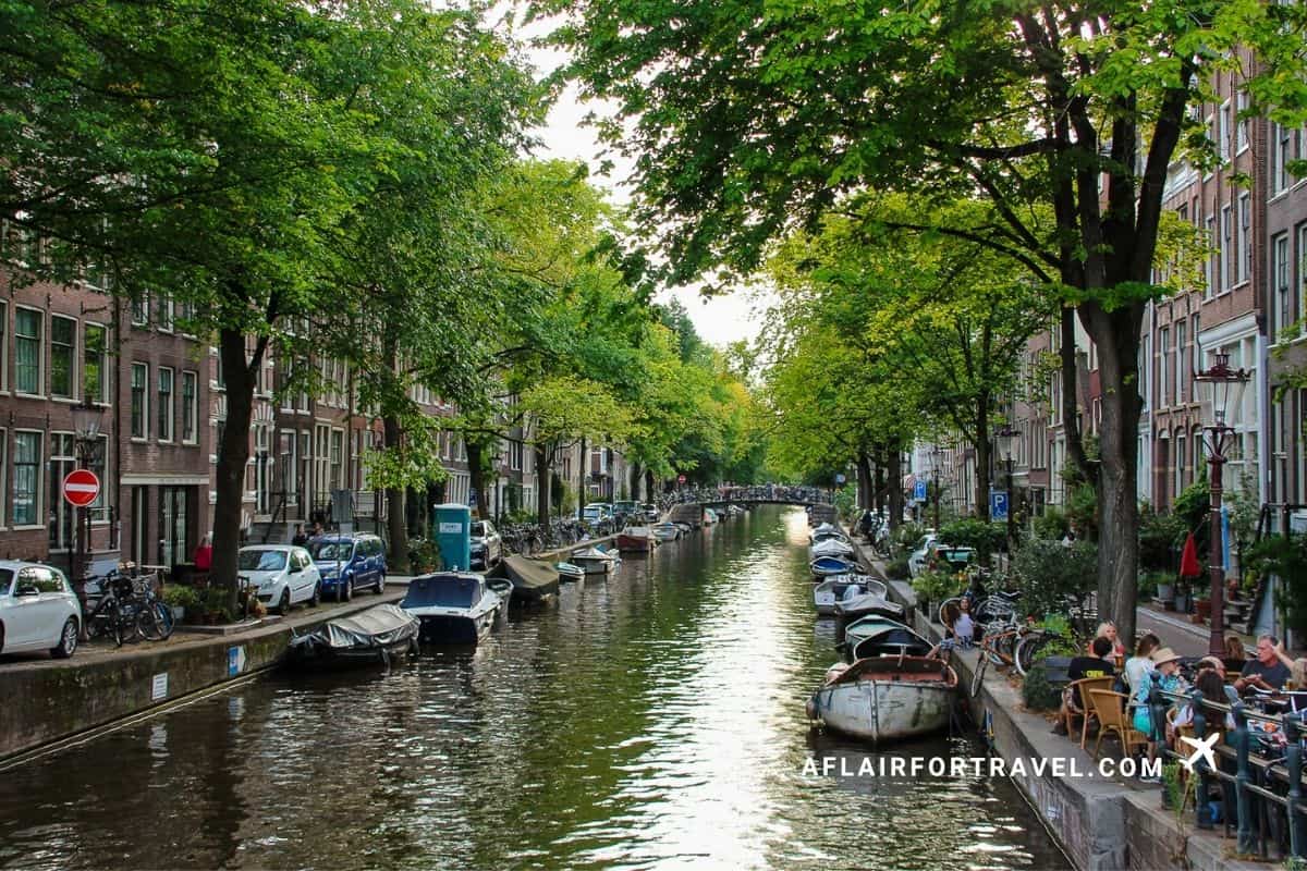 A quiet, tree-lined Amsterdam canal with houseboats and outdoor cafes along the water