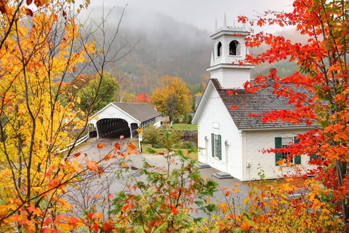 White church and covered bridge in the Great North Woods framed by fall foliage in a foggy mountain valley