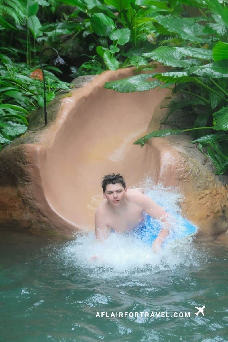 Teen boy sliding down a water slide surrounded by lush rainforest foliage at a hot springs resort in Costa Rica