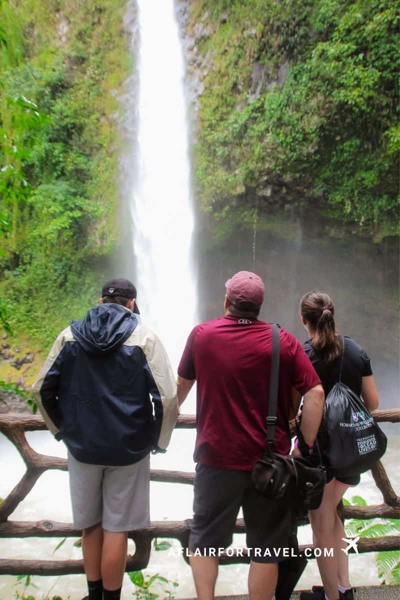 Family standing behind a fence admiring La Fortuna waterfall in Costa Rica; a popular tour to include when figuring out the cost of going to Costa Rica