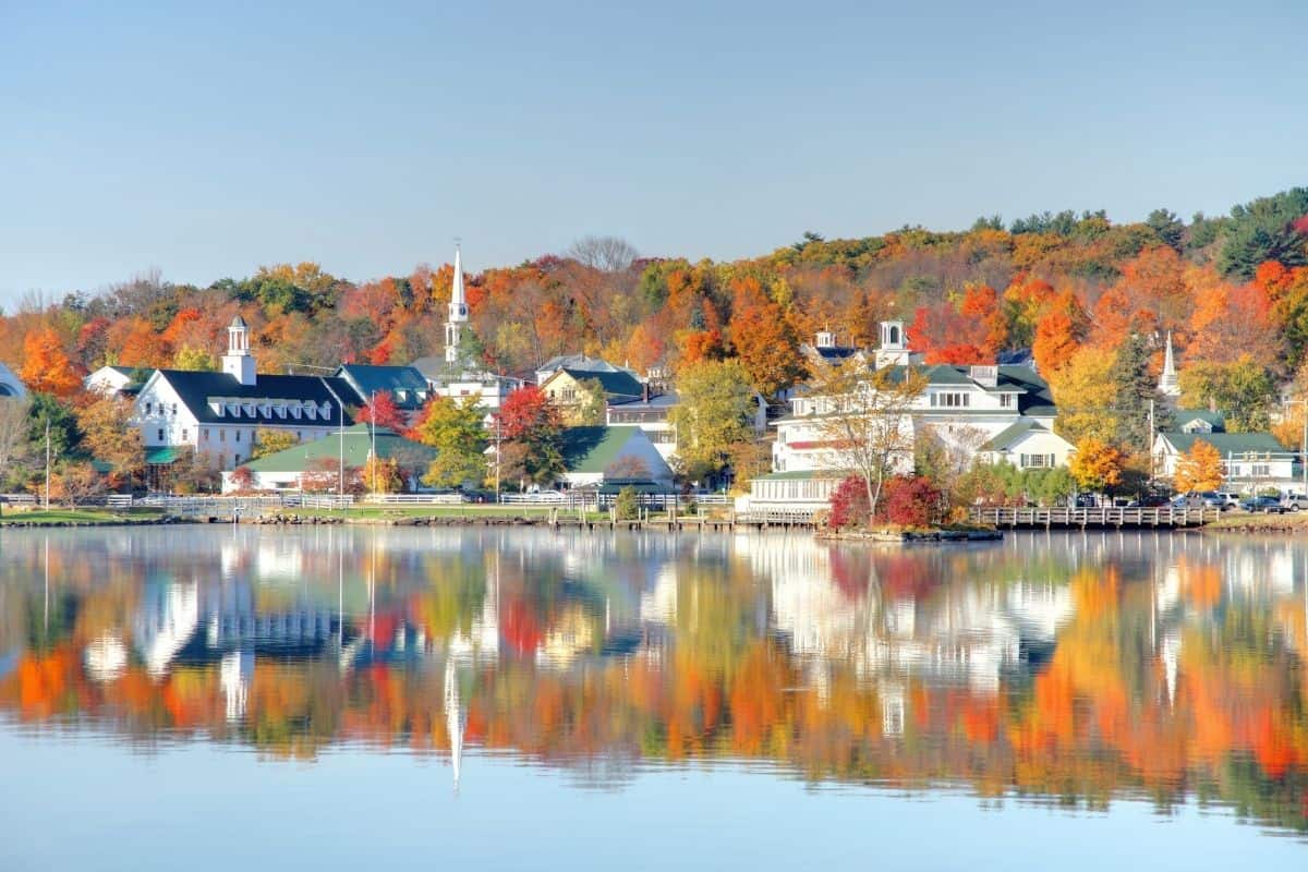New Hampshire fall scenery reflected in the water at Lake Winnipesaukee with colorful foliage surrounding a picturesque lakeside town.