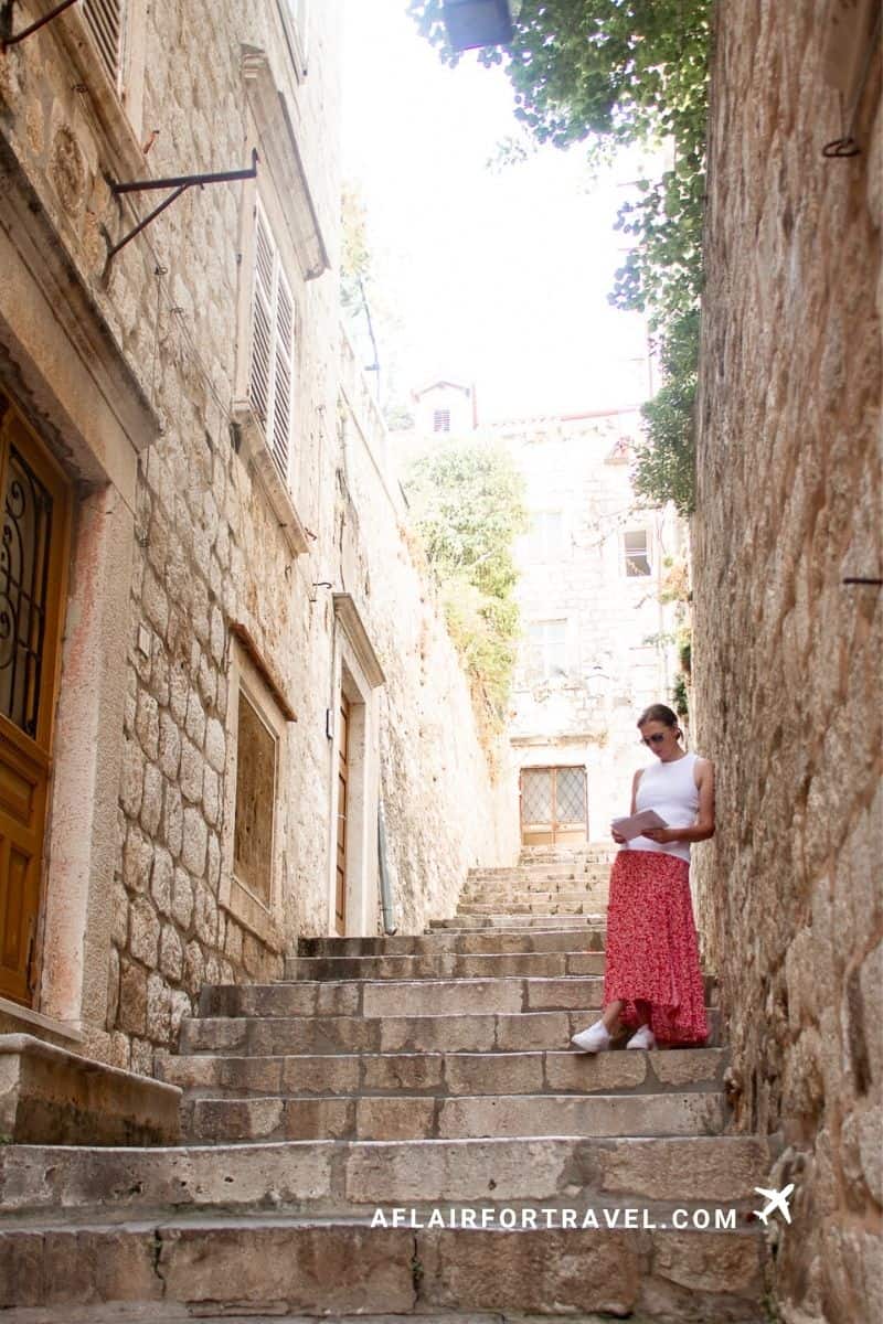 Woman reading a piece of paper while leaning against a wall along a narrow stone staircase in Dubrovnik's Old Town