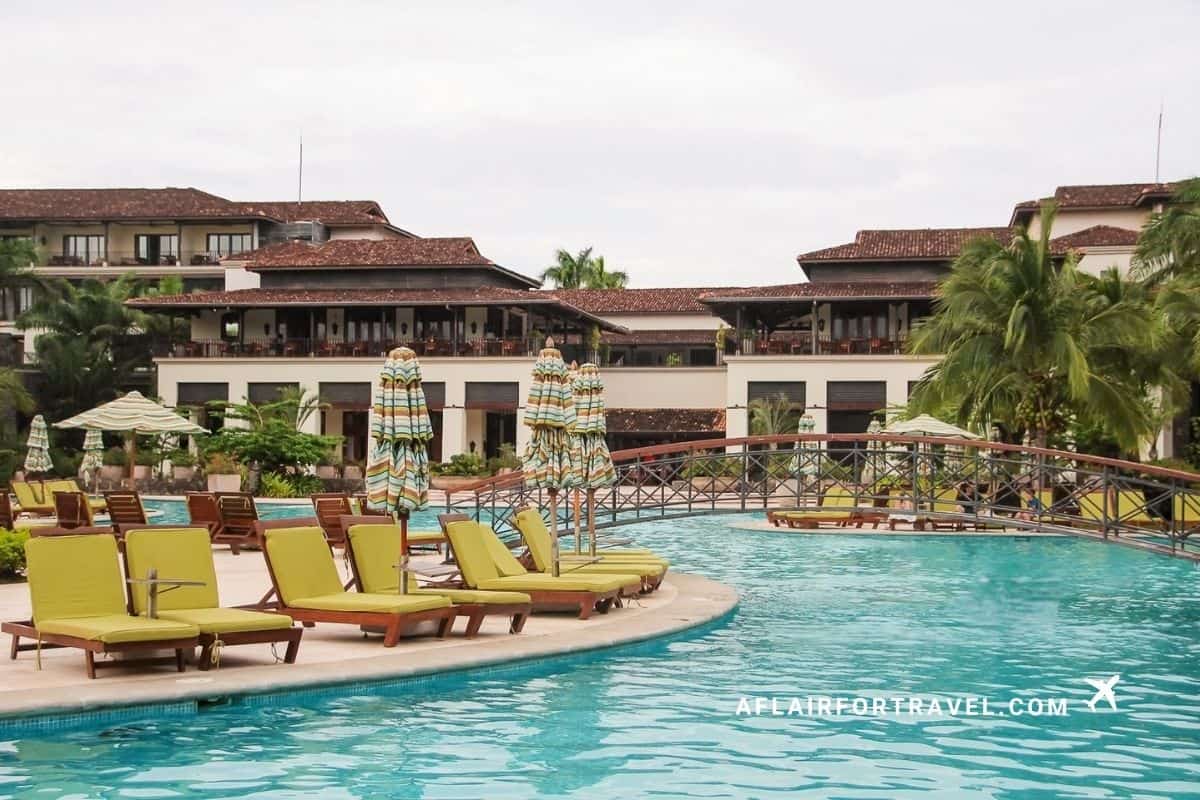 Large pool at a resort in Guanacaste with lounge chairs and striped umbrellas in front of hotel buildings