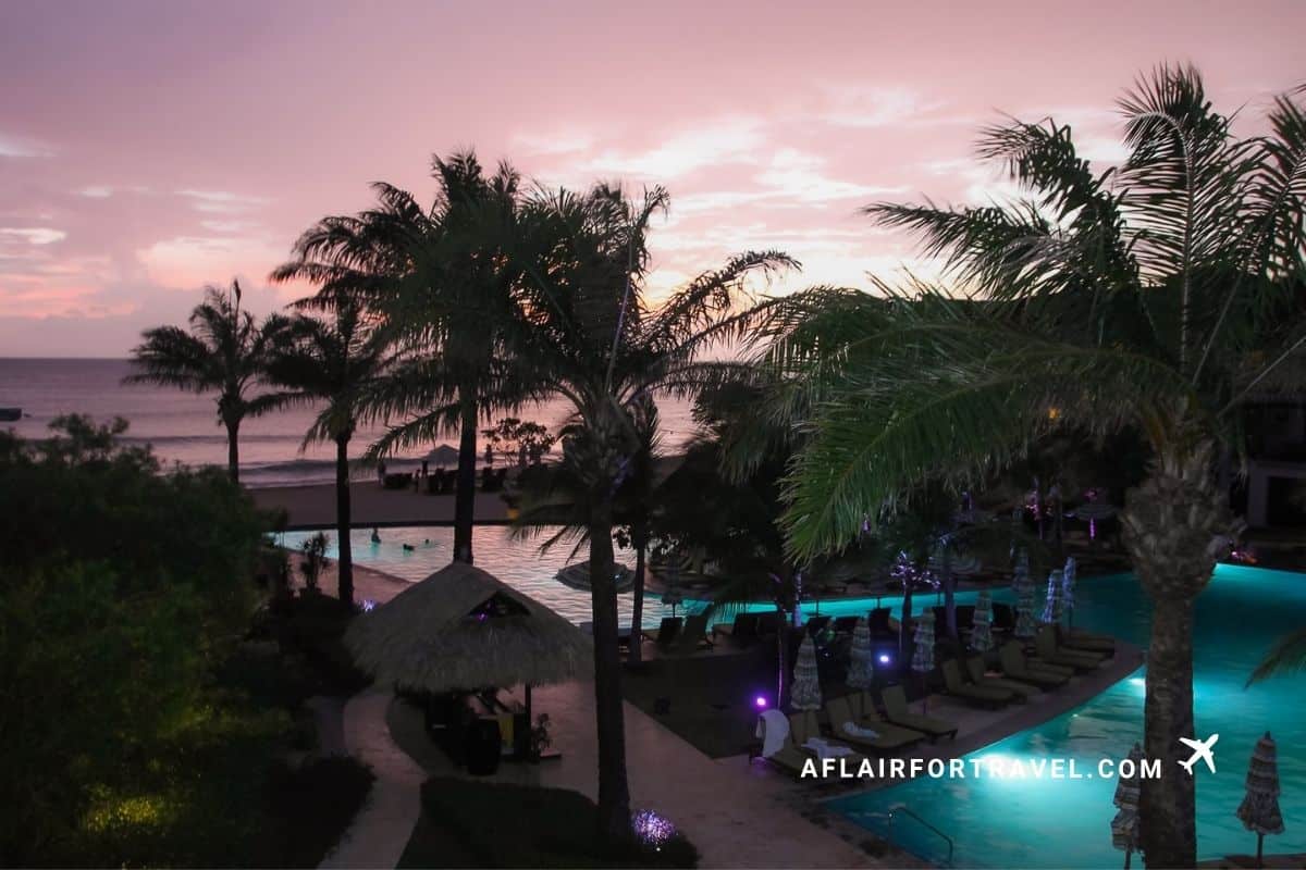 Resort pool illuminated at sunset with palm trees and beach in the distance